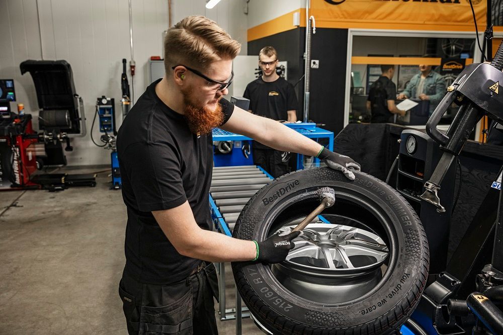 Mecánico montando un neumático en una máquina en un taller. Al fondo, otra persona y su equipo.