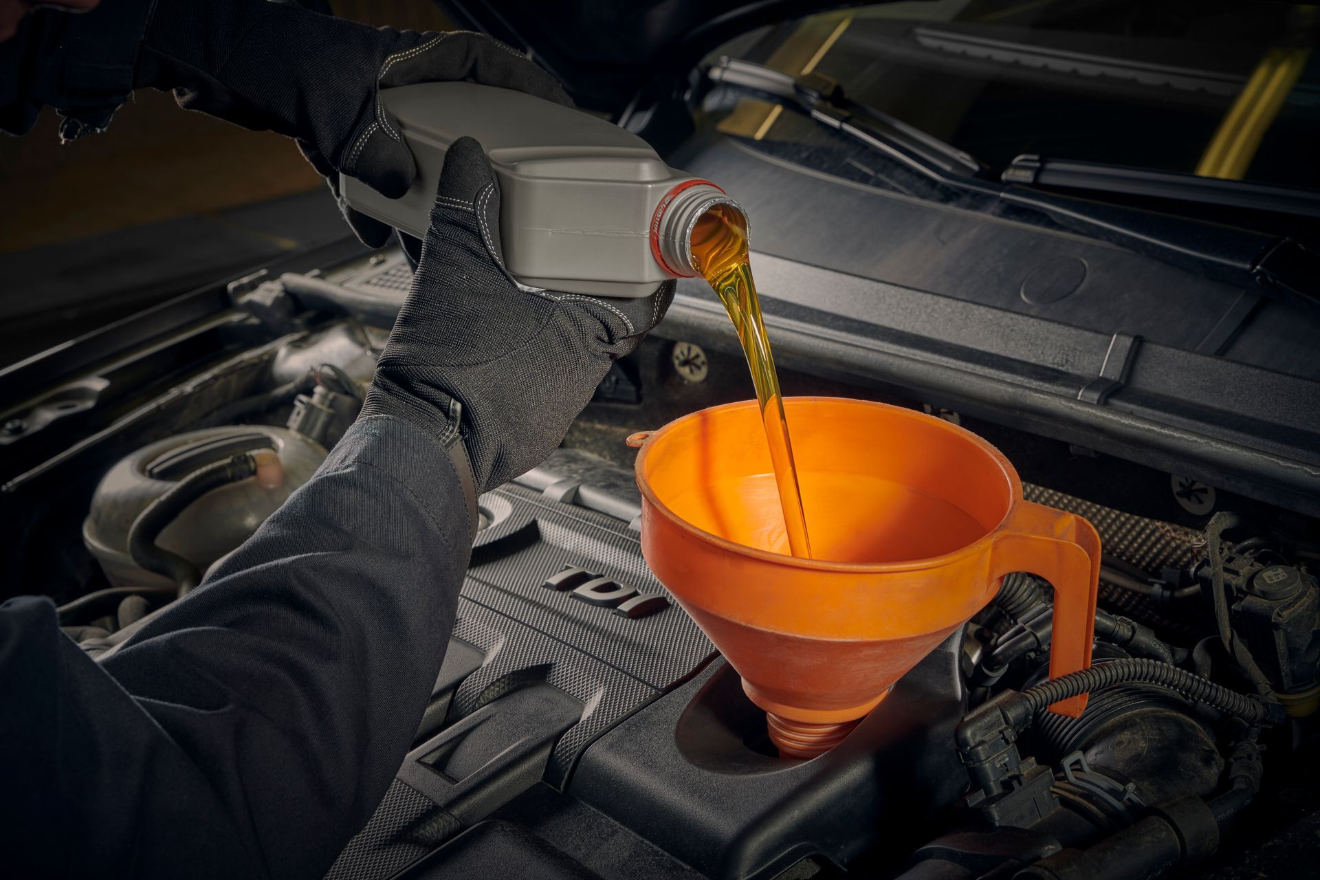 Persona vertiendo aceite en el motor de un coche con un embudo. Embudo naranja y guantes negros.