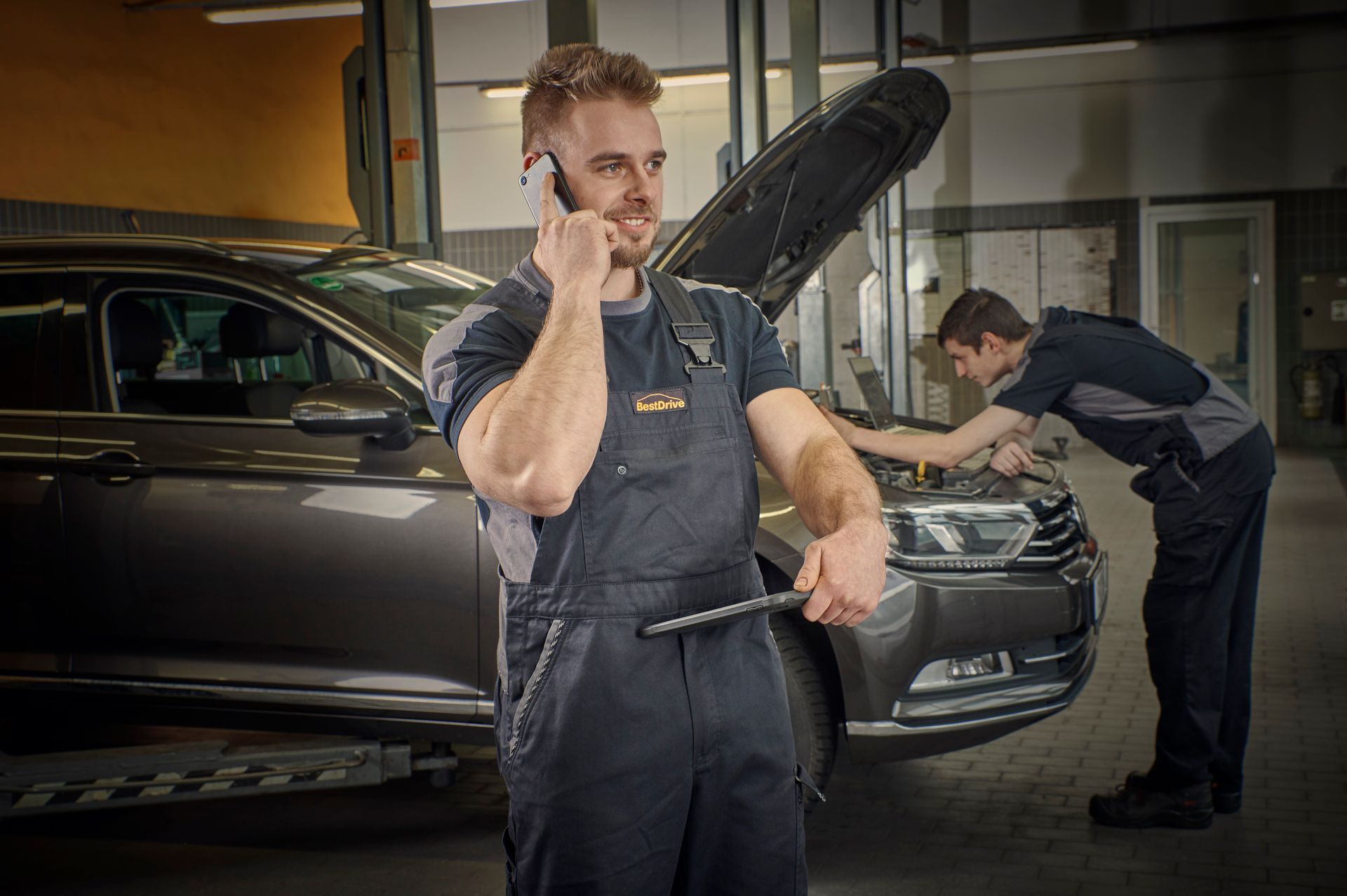 Mecánico hablando por teléfono, sosteniendo una herramienta, en un taller de reparación de automóviles con otro mecánico trabajando en el automóvil.