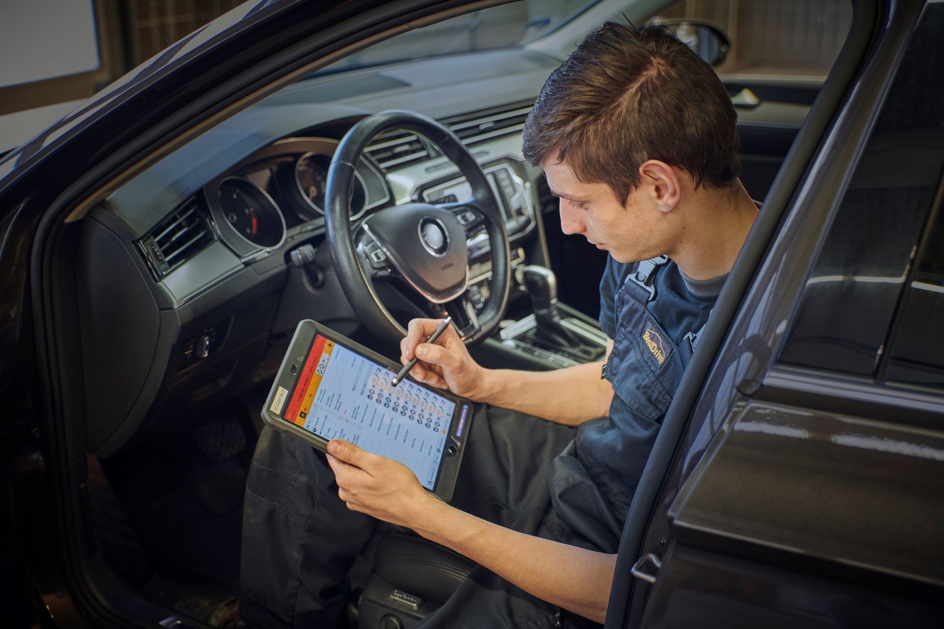 Mecánico en coche, usando tableta para diagnóstico. Interior oscuro, coche negro, mirando la pantalla.