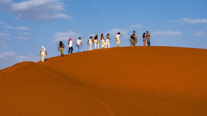 Grupo de personas caminando sobre una gran duna de arena naranja bajo un cielo azul.