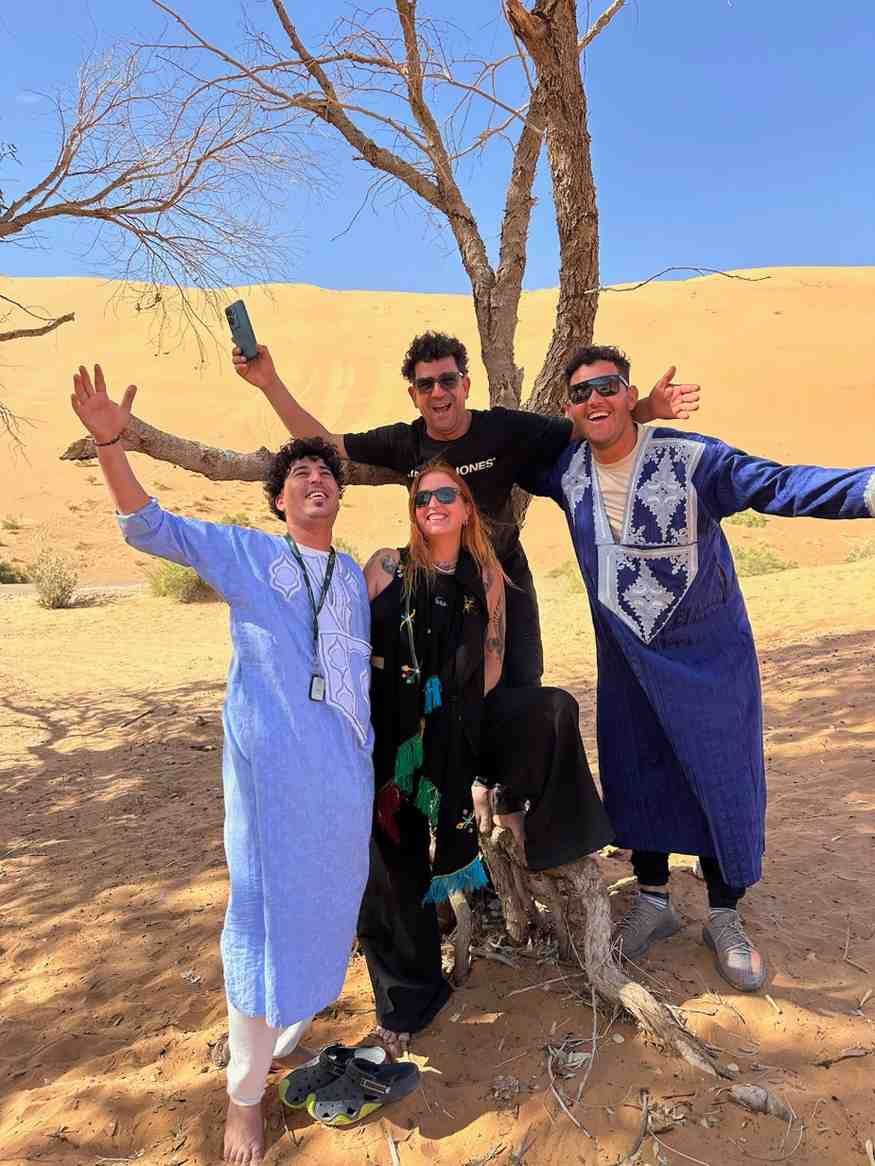 Cuatro personas posan bajo un árbol en el desierto. Una sostiene un teléfono y las demás tienen los brazos en alto.