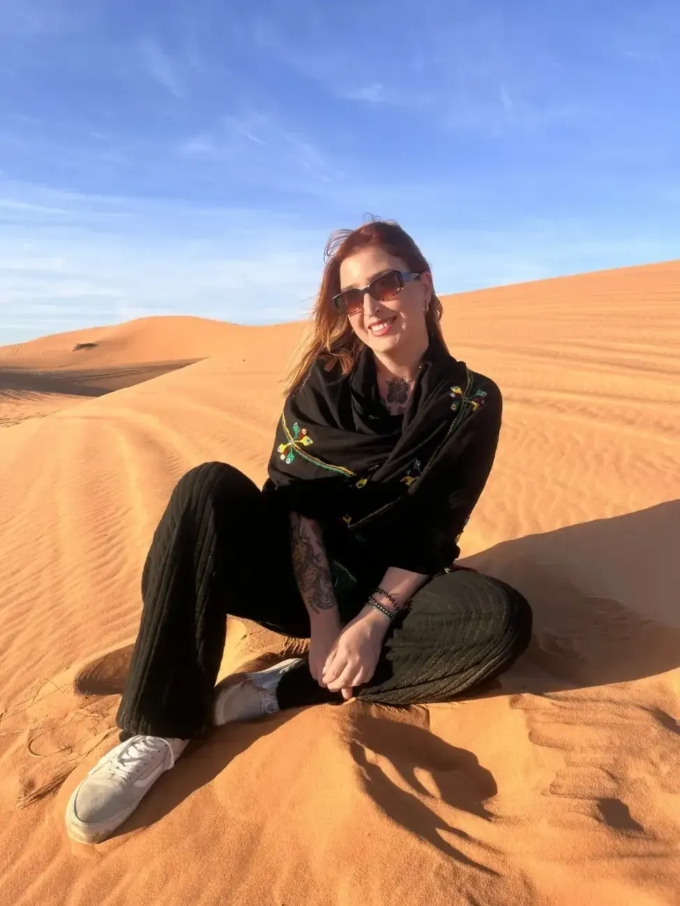 Mujer sentada en una duna de arena, vestida con ropa negra y gafas de sol, sonriendo. Paisaje desértico.