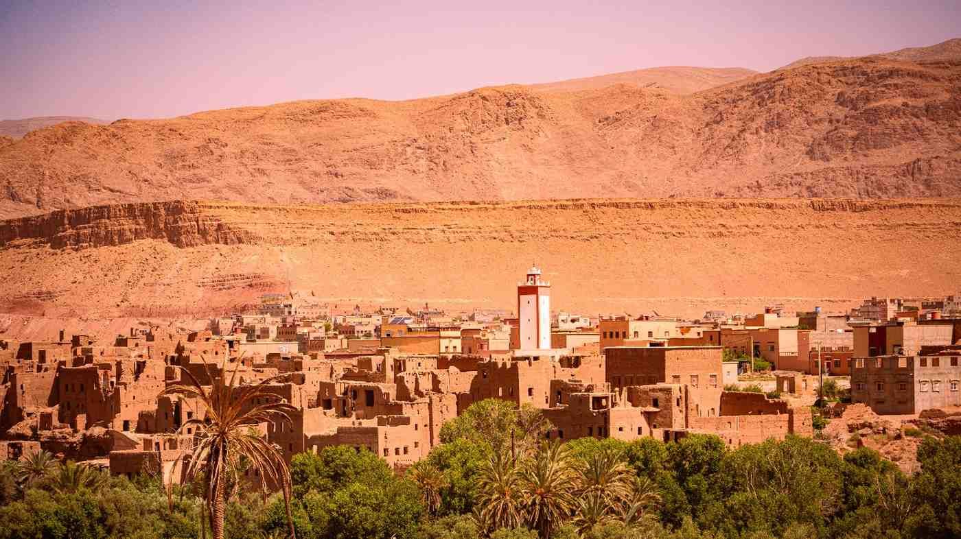 Ciudad del desierto con edificios de adobe y un minarete, bajo una cadena montañosa de color canela.