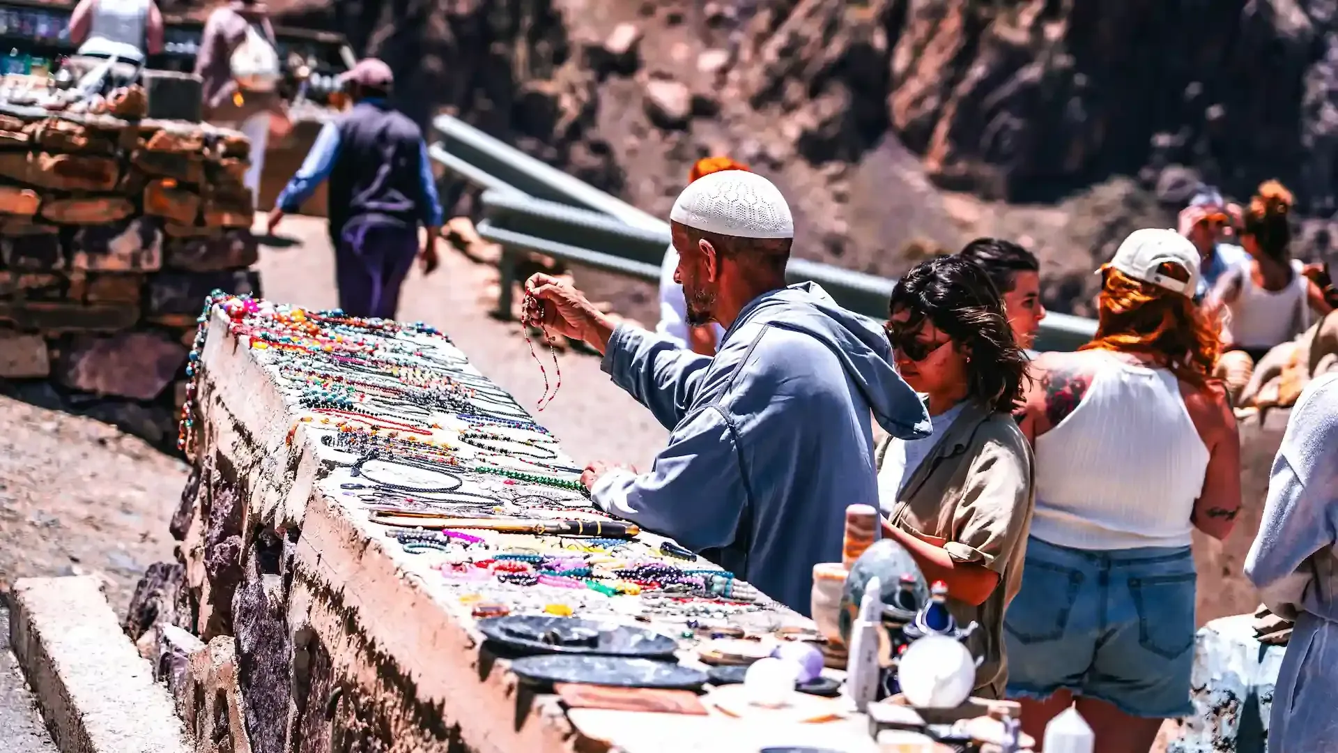 Hombre vendiendo joyas en un mercado al aire libre. Los compradores miran. Día soleado con fondo rocoso.