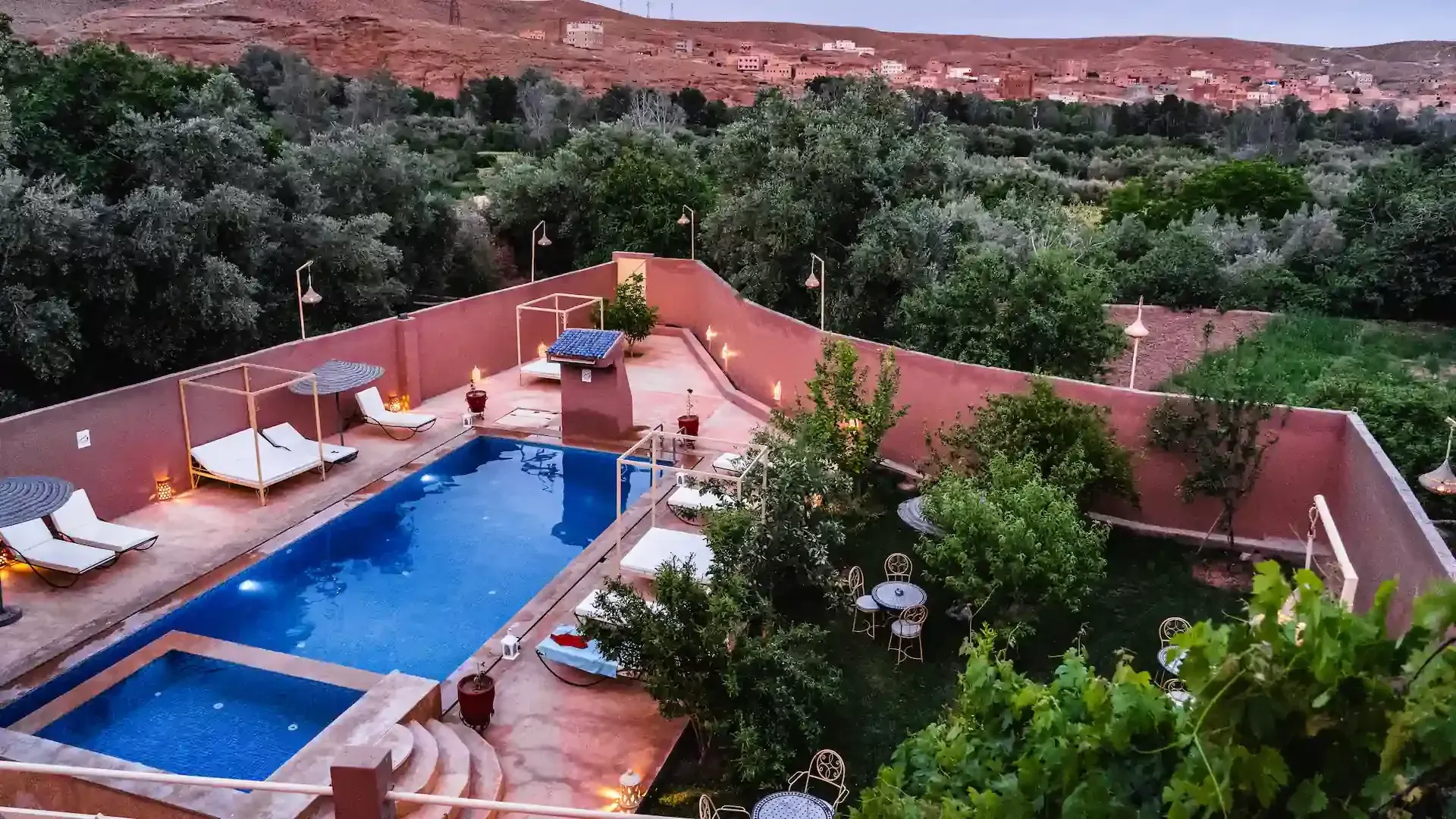 Zona de piscina con tumbonas, rodeada de paredes rojas y exuberante vegetación, en un entorno desértico.