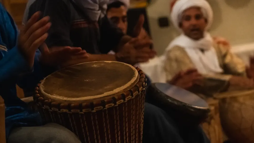Gente tocando tambores en un interior; una mano aplaude. Iluminación cálida.