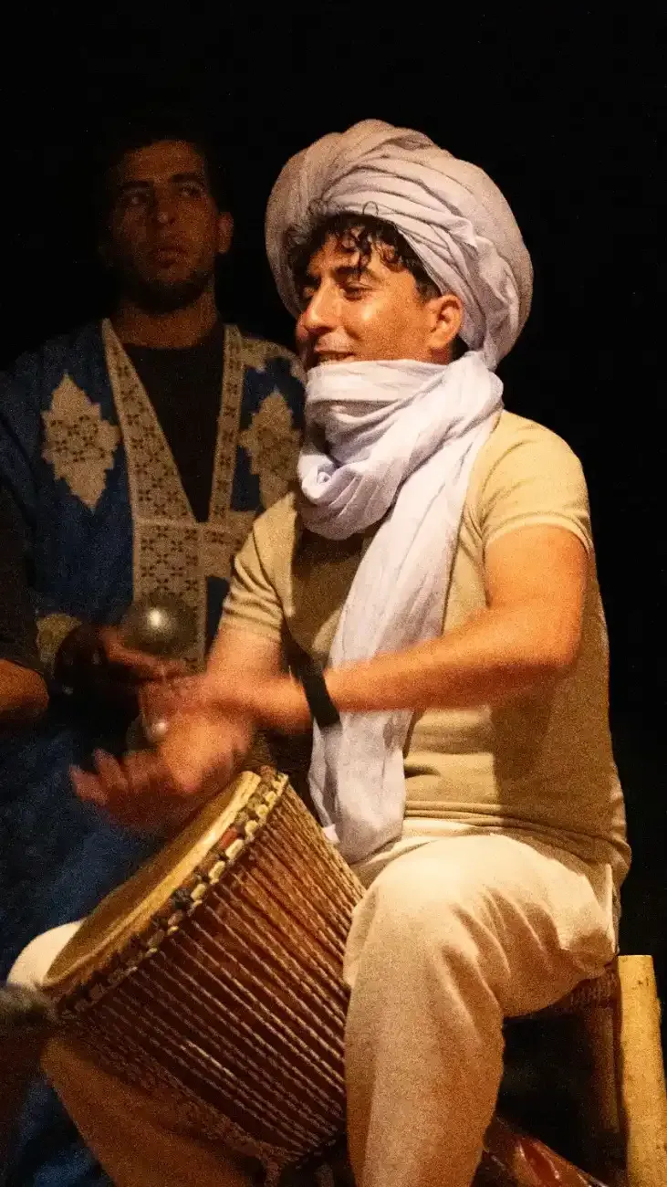 Hombre tocando un tambor, con turbante y bufanda, con otro hombre detrás de él.