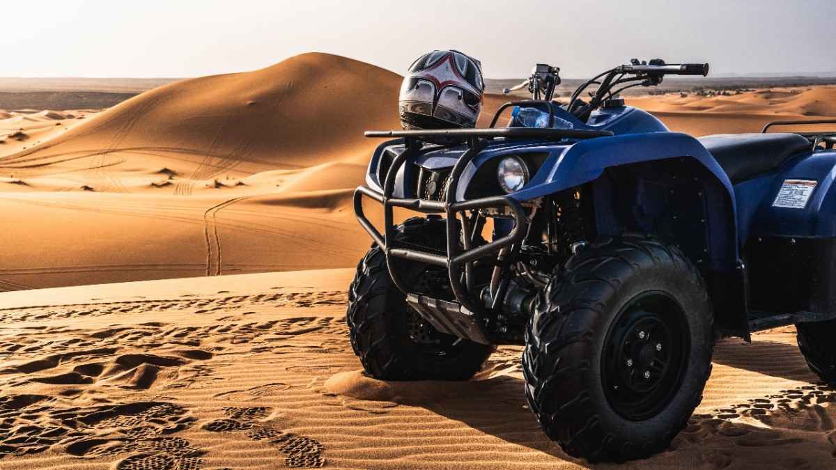 Vehículo todo terreno azul estacionado en un desierto arenoso, con el casco en el portaequipaje delantero.
