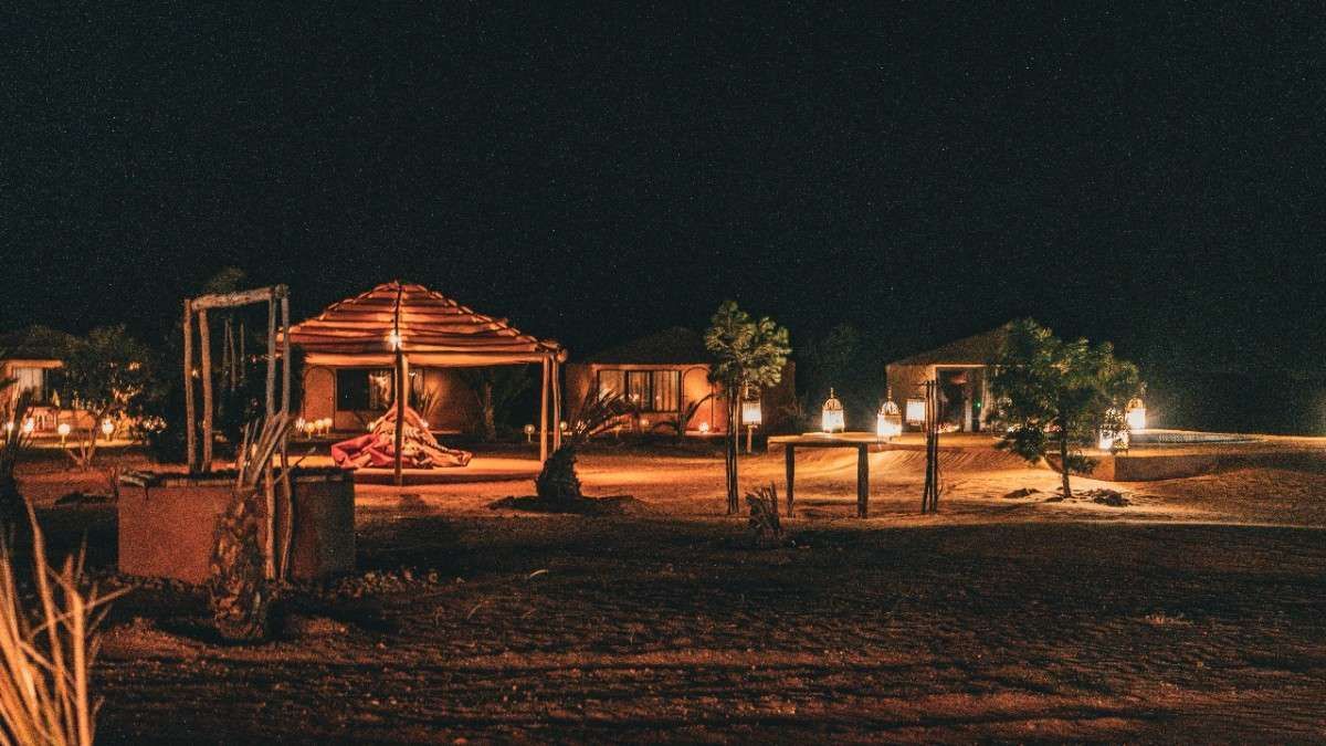 Campamento en el desierto por la noche con cabañas iluminadas y árboles bajo un cielo estrellado.