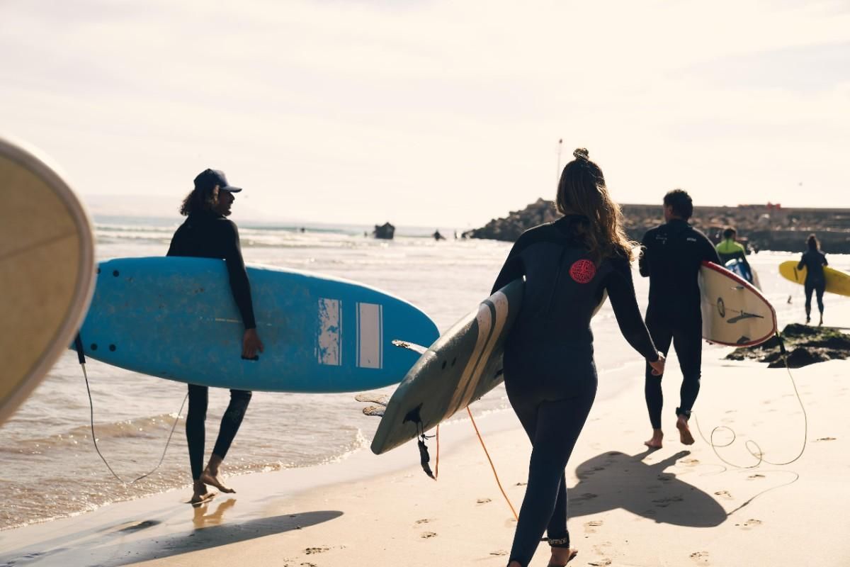 Surfistas con trajes de neopreno llevan tablas a lo largo de una playa de arena hacia el océano en un día soleado.