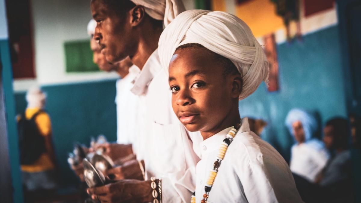 Un niño con turbante blanco mira a la cámara. Otros, vestidos de blanco, están cerca.