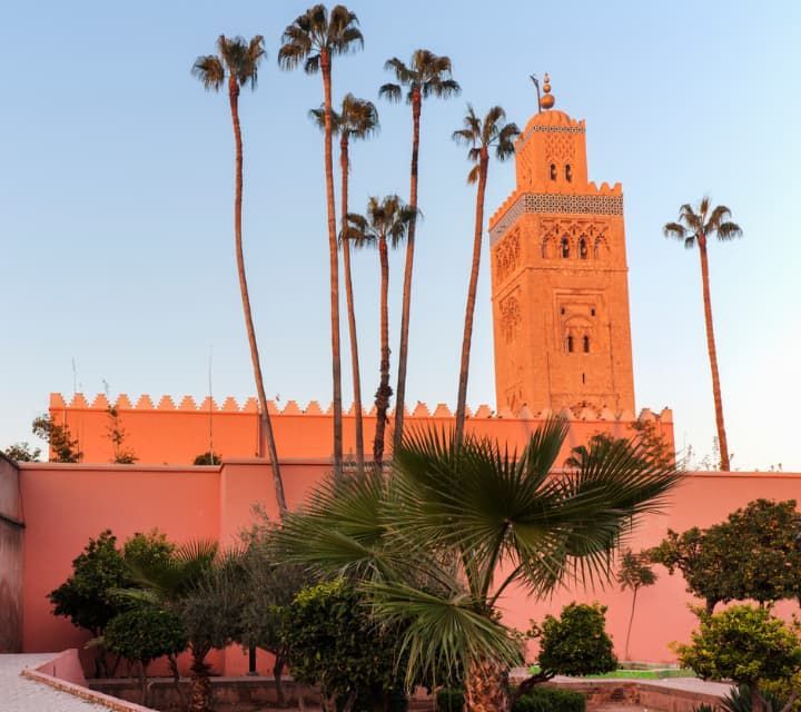 Alto minarete y muro de la Mezquita Koutoubia en Marrakech, Marruecos, con palmeras contra un cielo azul.
