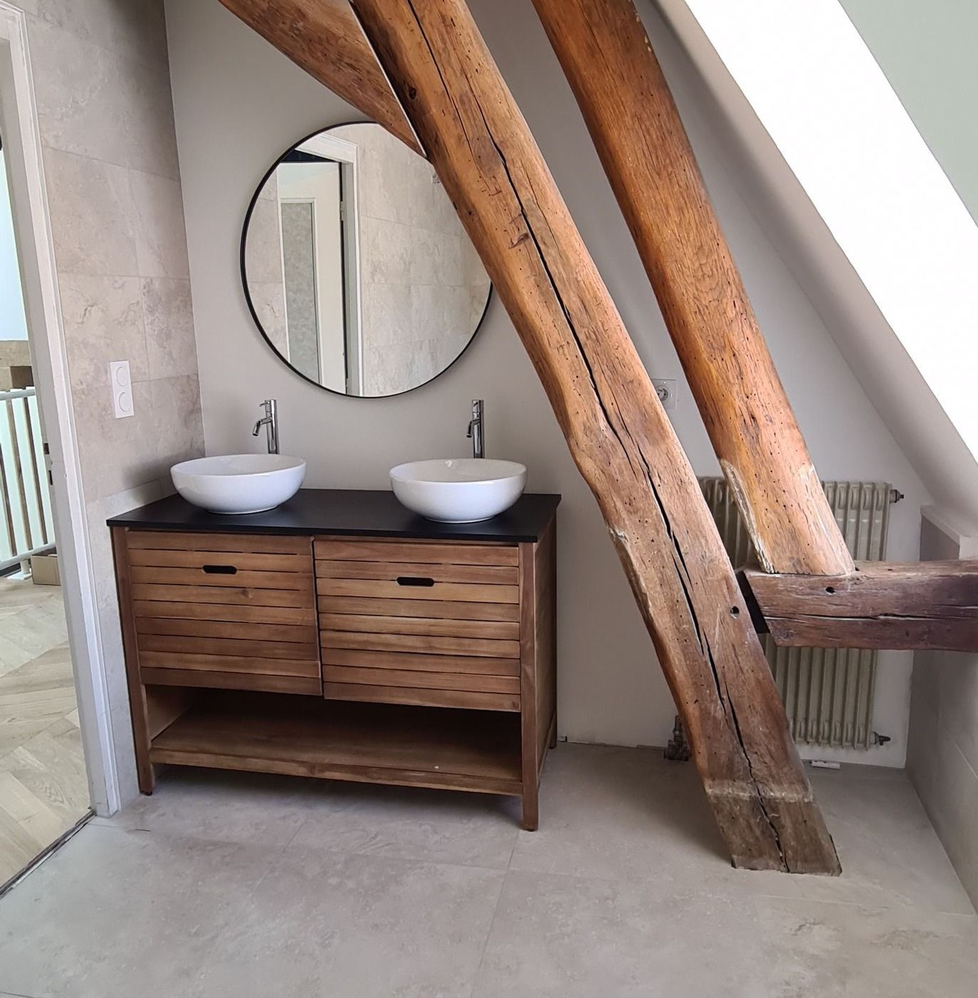 Salle de bains avec poutres apparentes, double vasque, miroir rond et meuble-lavabo en bois.