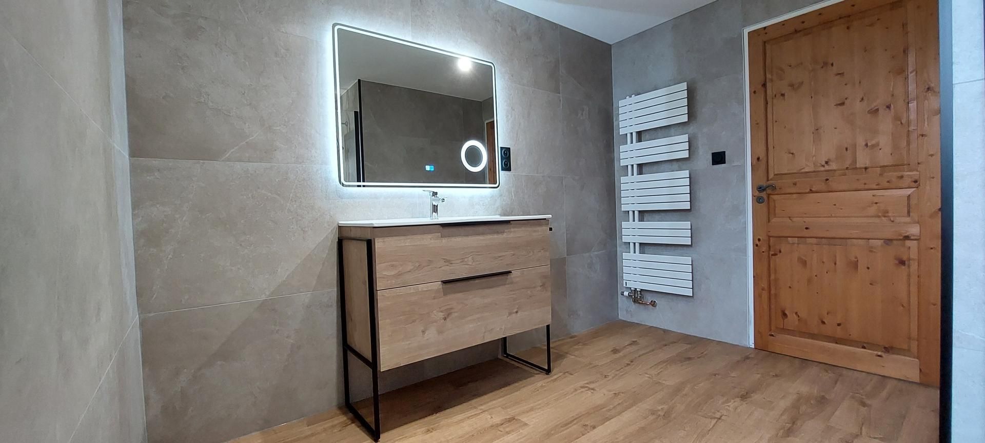 Salle de bains avec meuble lavabo en bois, miroir éclairé, sèche-serviettes et porte en bois.