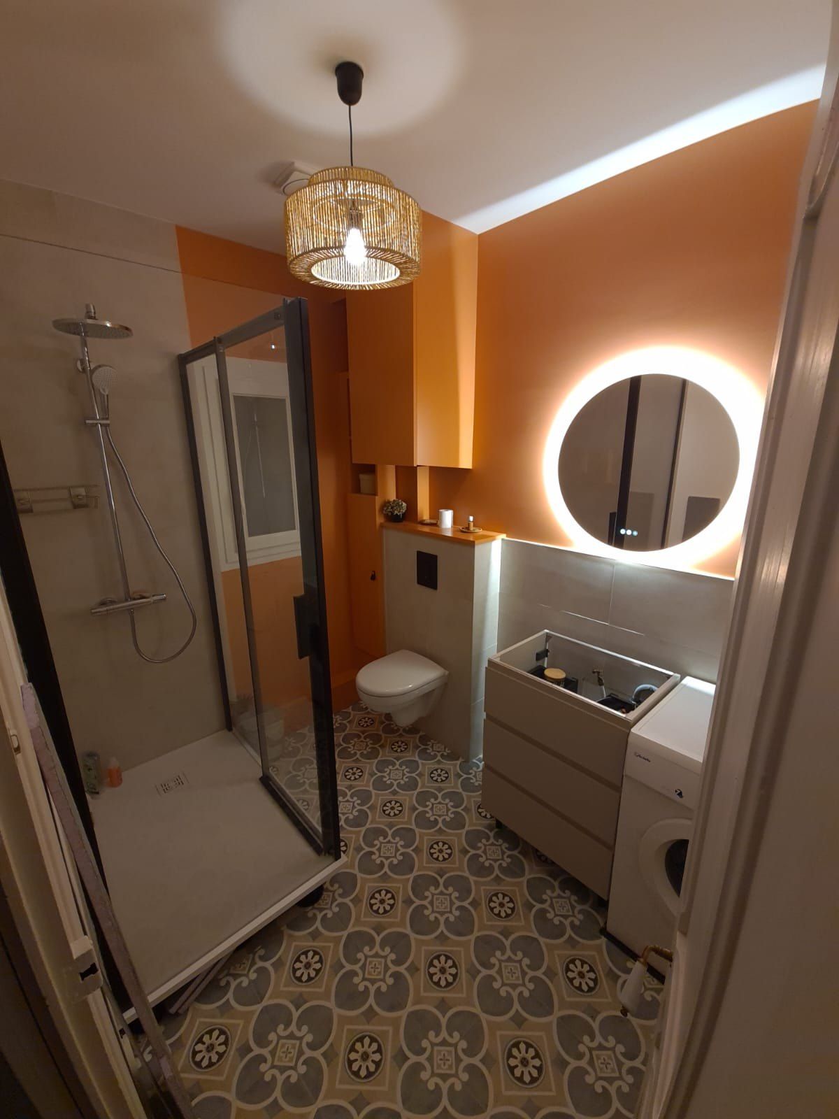 Petite salle de bains aux murs orange, sol à motifs floraux, douche et miroir rond rétroéclairé.