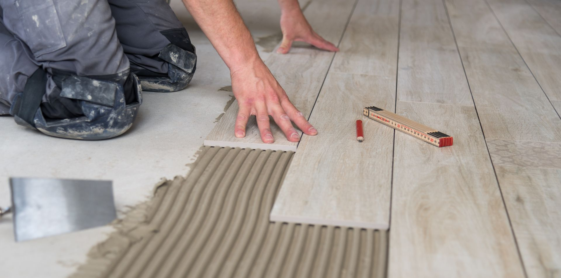 Une personne pose des carreaux de parquet en appliquant de la colle. Elle est agenouillée et utilise une truelle et un outil de mesure.
