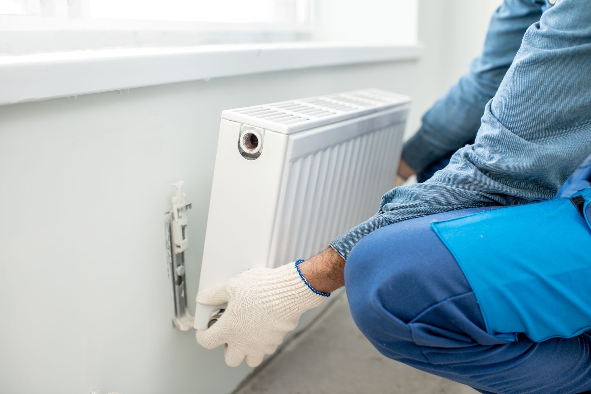 Une personne installe un radiateur blanc près d'une fenêtre. Elle porte des gants et une combinaison de travail bleue.