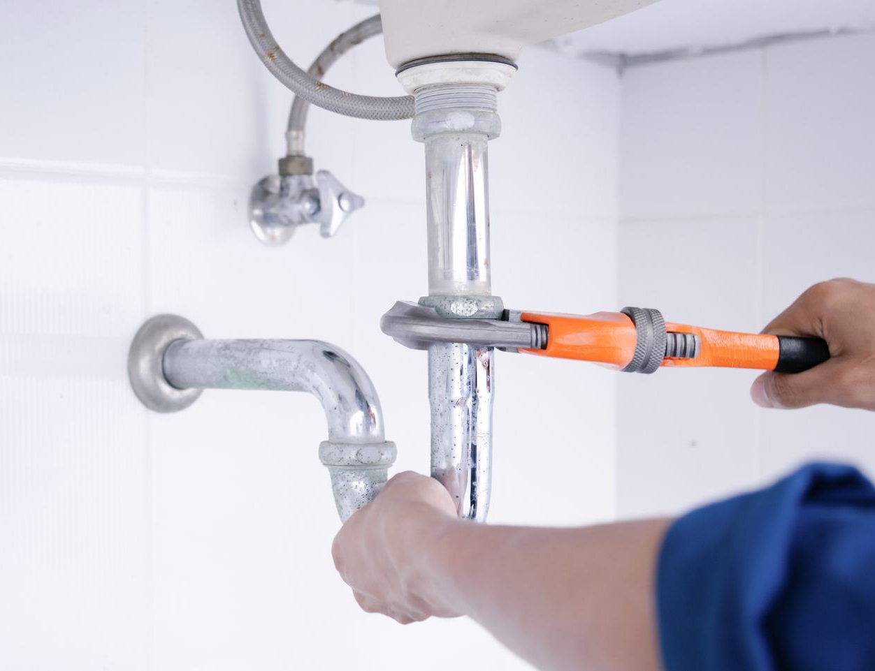 Une personne utilise une clé orange pour serrer un tuyau sous un évier blanc dans une pièce carrelée.