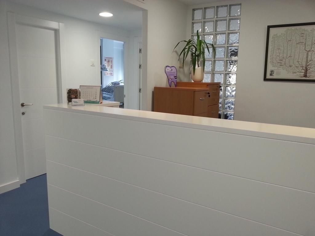 Mostrador de recepción blanco en un consultorio médico. Una planta y una obra de arte se encuentran sobre un armario. Se ve un pasillo.