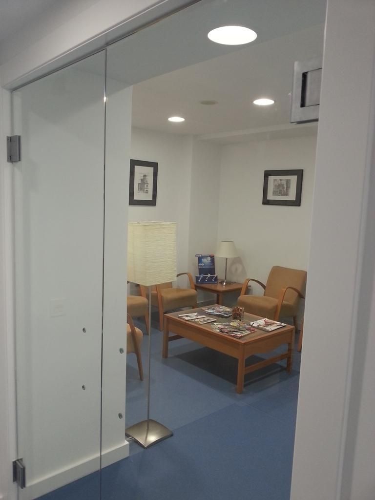 Puerta de cristal que da a una sala de espera con sillas, lámpara y mesa de centro. Suelo azul, paredes blancas.
