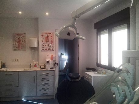 Interior del consultorio dental: silla de dentista, luz de techo, armarios, ventana con persianas, historiales médicos en la pared.