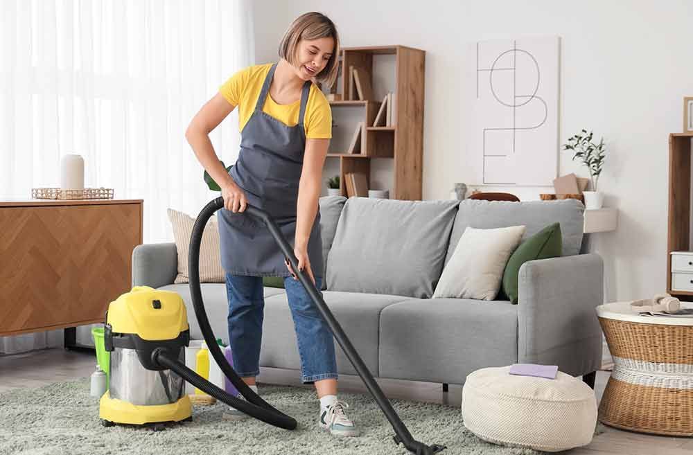 Eine Frau saugt lächelnd den Teppichboden ihres Wohnzimmers. Sie trägt eine Schürze. Der gelbe Staubsauger ist zu sehen.