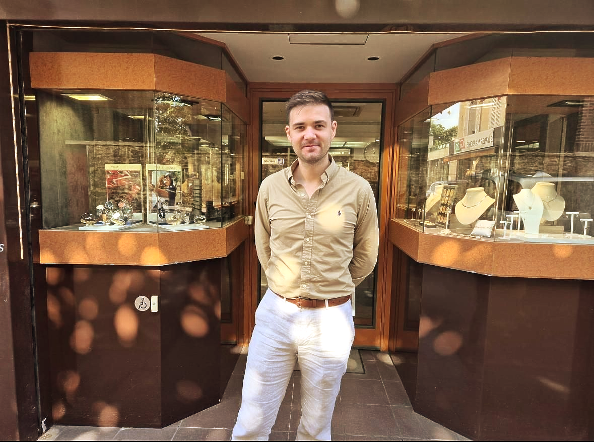 Homme debout devant une bijouterie, les bras derrière le dos. Des bijoux sont exposés dans des vitrines.