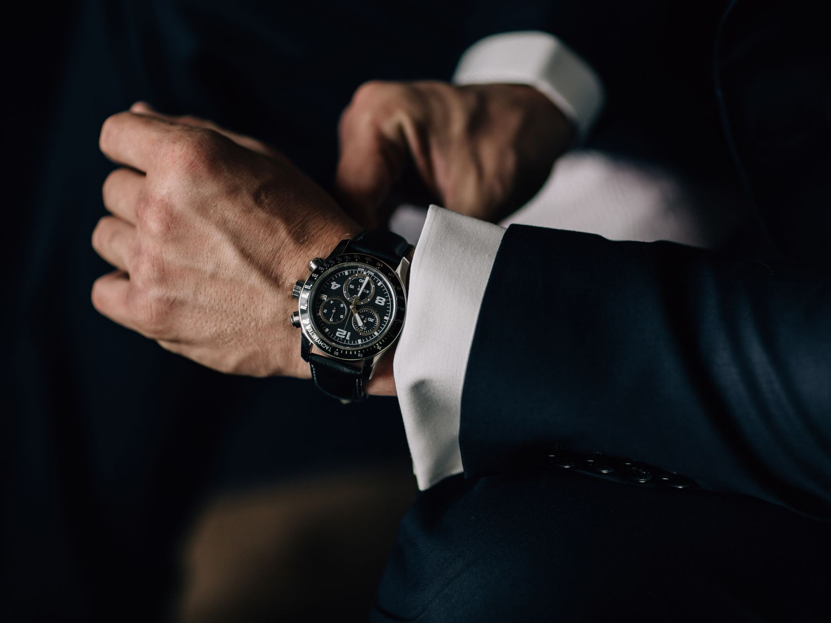 Homme en costume ajustant une montre-bracelet noire. Fond sombre, poignet de chemise blanc visible.