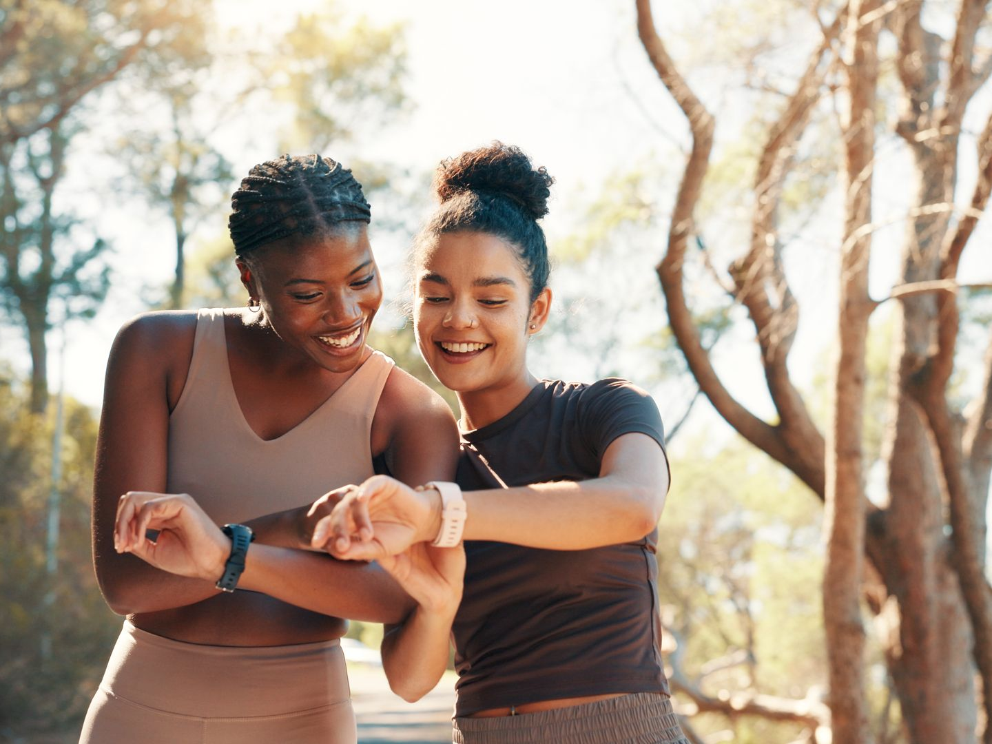 Deux femmes sourient en regardant leurs montres connectées, dehors. Arbres et soleil en arrière-plan.
