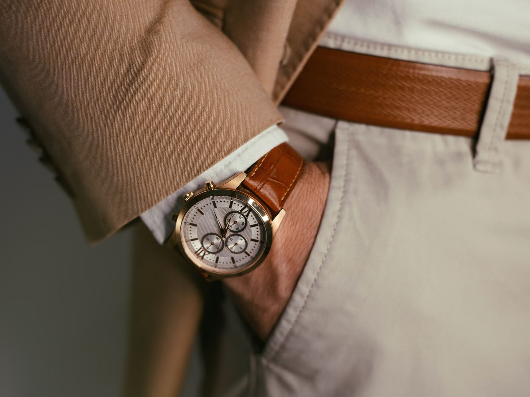 Homme portant une montre en or avec bracelet en cuir marron, bras dans la poche d'un pantalon kaki, veste beige.