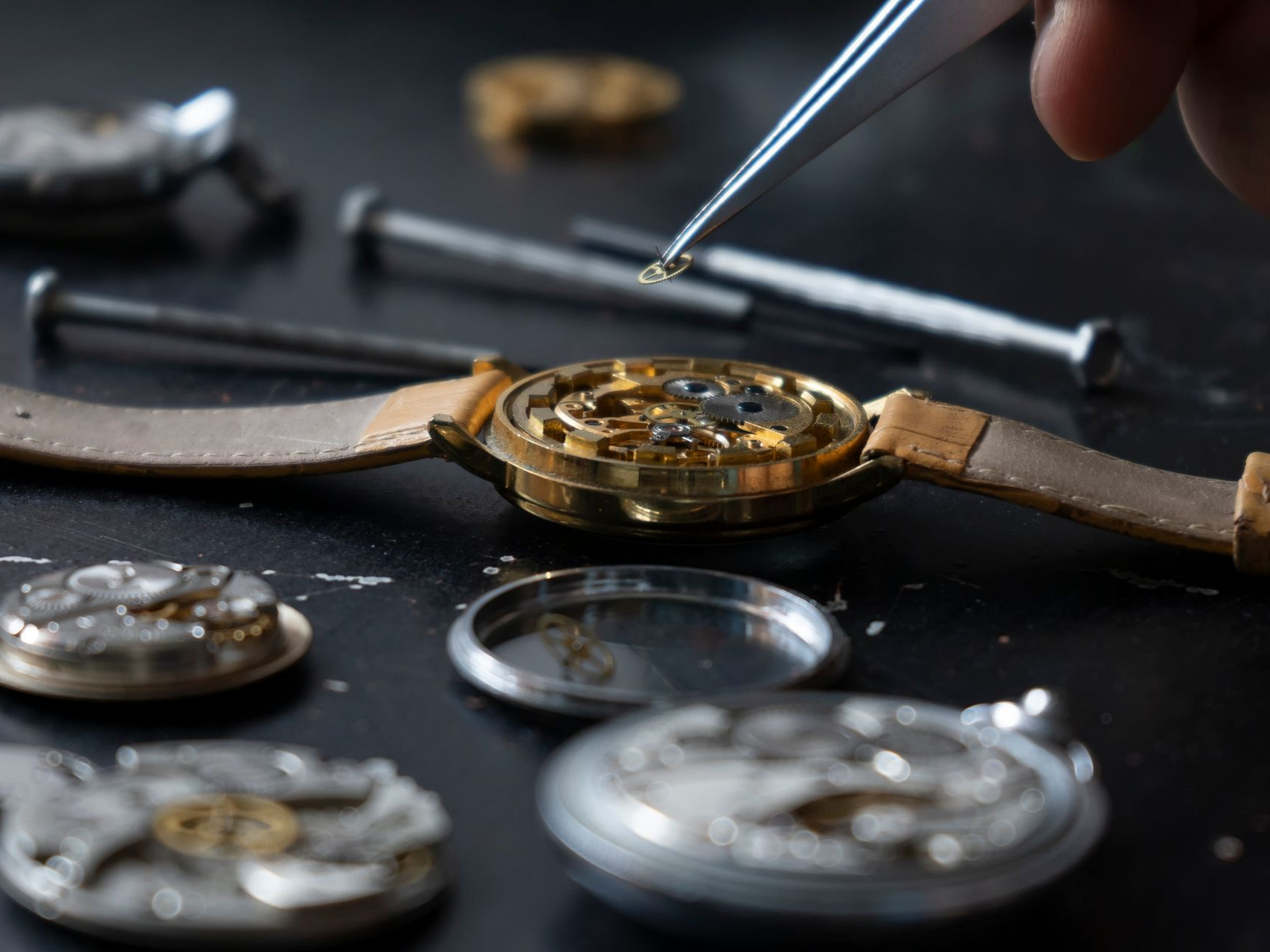 Horloger réparant une montre en or démontée avec des pincettes, petites pièces sur une surface noire.