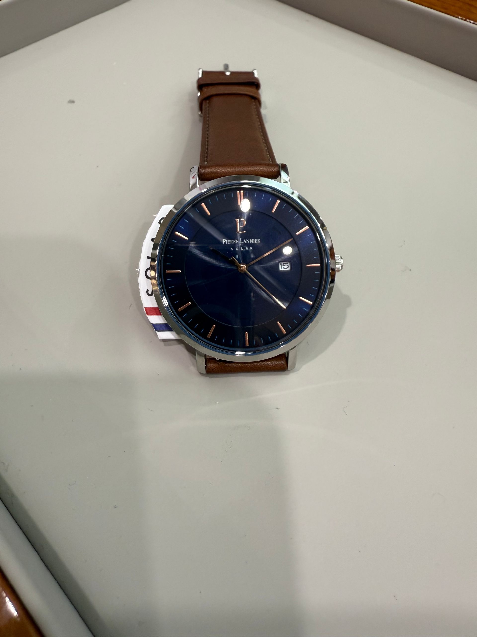 Homme portant une montre en or avec bracelet en cuir marron, bras dans la poche d'un pantalon kaki, veste beige.