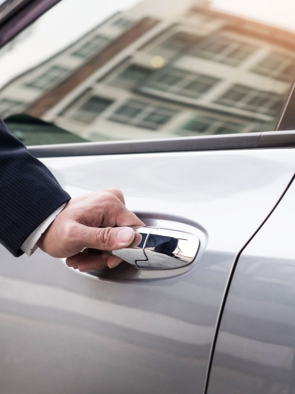 Une main ouvrant une poignée de portières de voiture argentée, avec le reflet d'un immeuble.