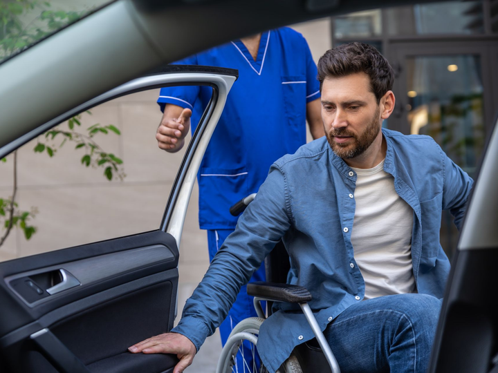 Un homme en fauteuil roulant est aidé à monter dans une voiture par une personne en blouse bleue.