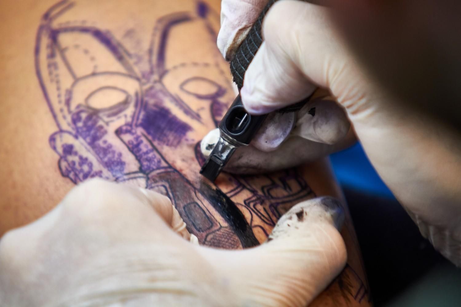 El brazo de una persona siendo tatuado por alguien que usa guantes, con un diseño morado parcialmente terminado.