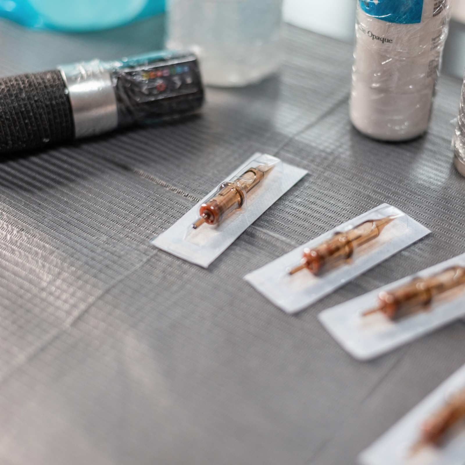 Agujas de tatuaje en embalaje estéril sobre una superficie gris, con un mango de máquina de tatuaje en el fondo.