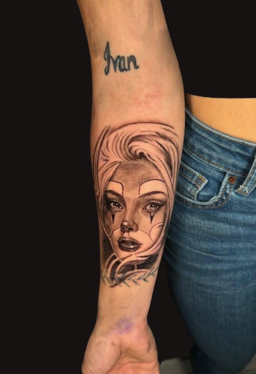 Tatuaje en el antebrazo del rostro de una mujer con cabello claro y rasgos oscuros. La palabra 
