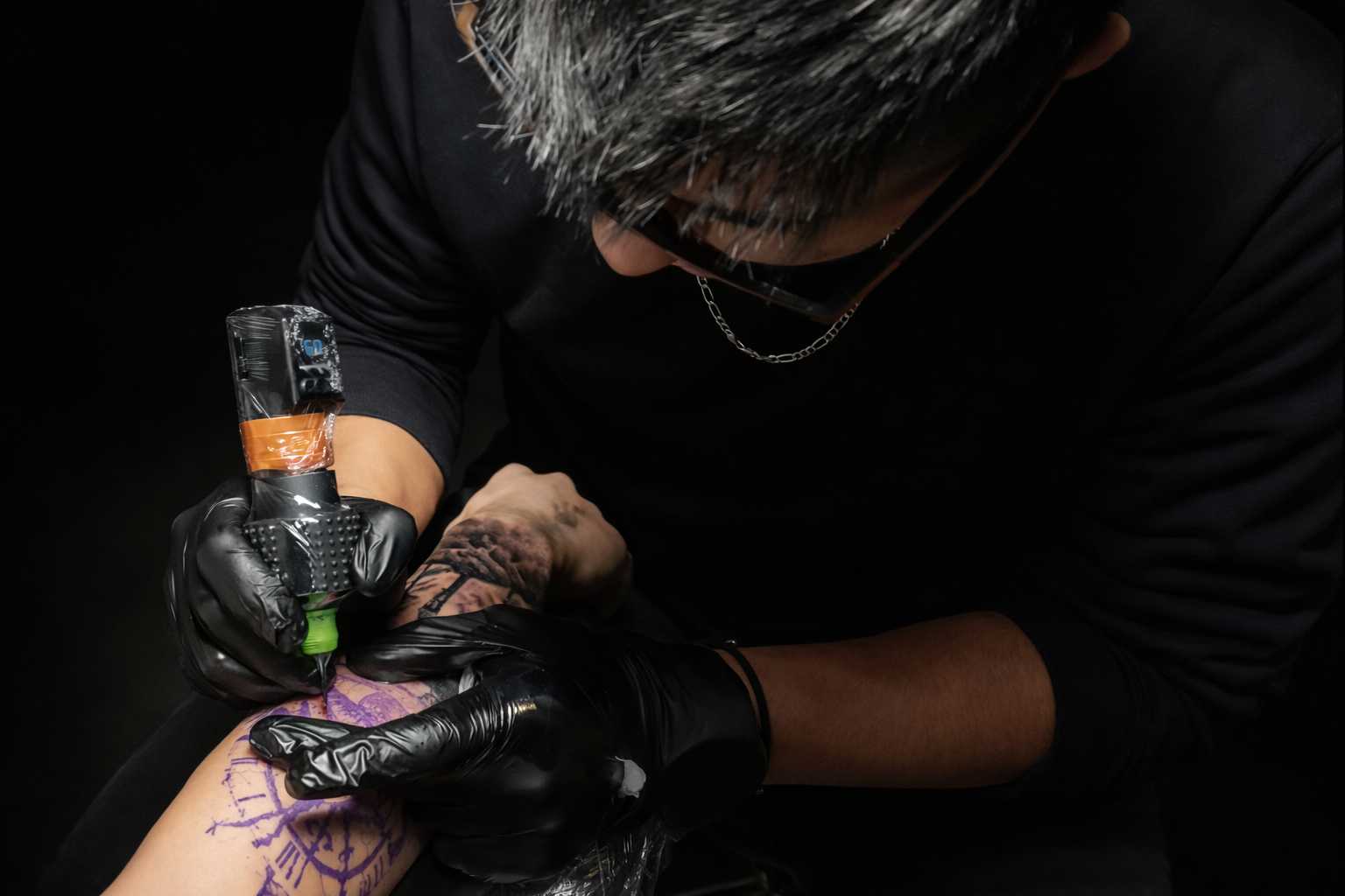 Artista del tatuaje con guantes negros vertiendo tinta en vasos de color, herramientas y botellas sobre una superficie blanca.
