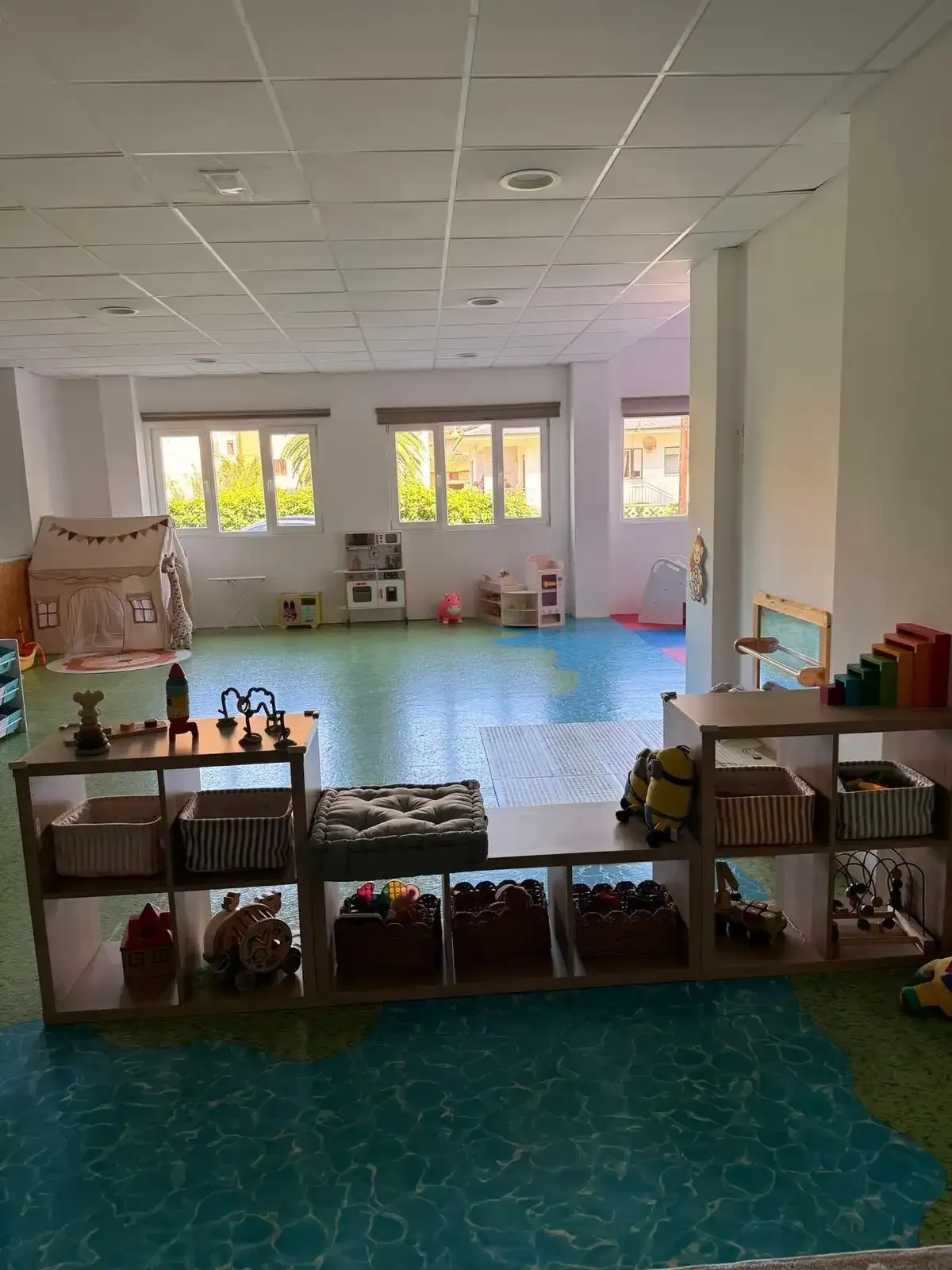 Aula vacía con suelo azul, estanterías llenas de juguetes y grandes ventanas al fondo.
