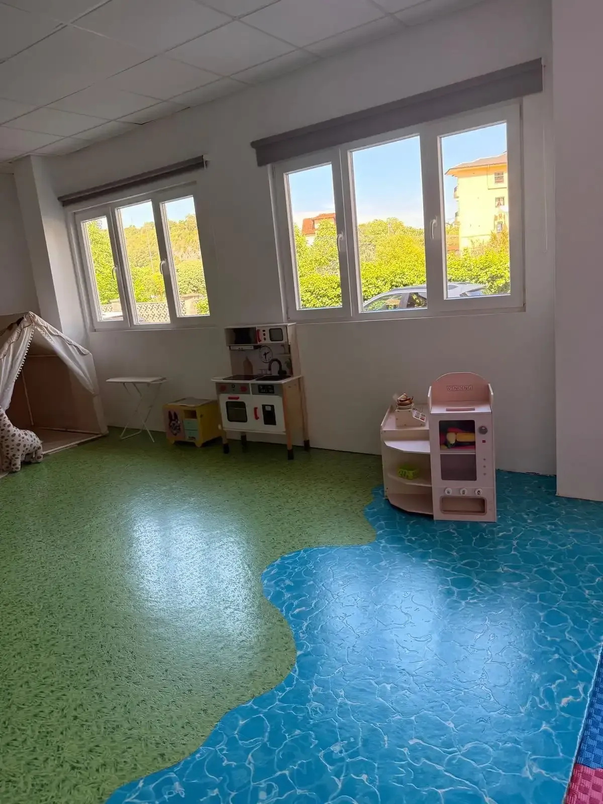 Habitación luminosa con suelo estampado en verde y azul, ventanas, estanterías y una pequeña cocinita de juguete.