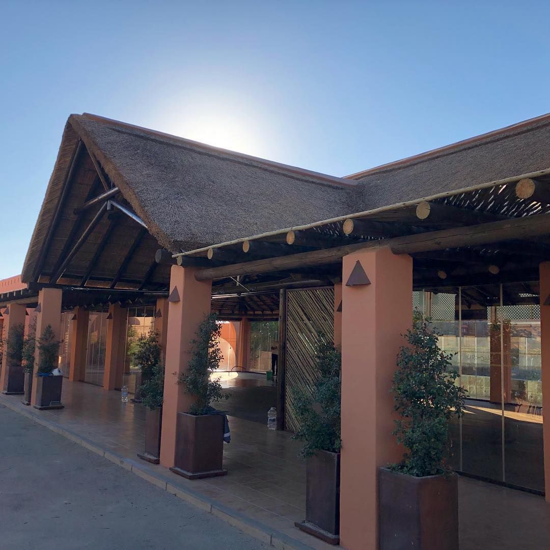 Edificio de terracota con techo de paja y pilares, plantas en macetas, cielo despejado.