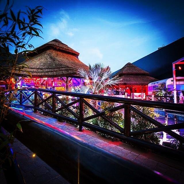 Bar con techo de paja iluminado y puente de madera por la noche.