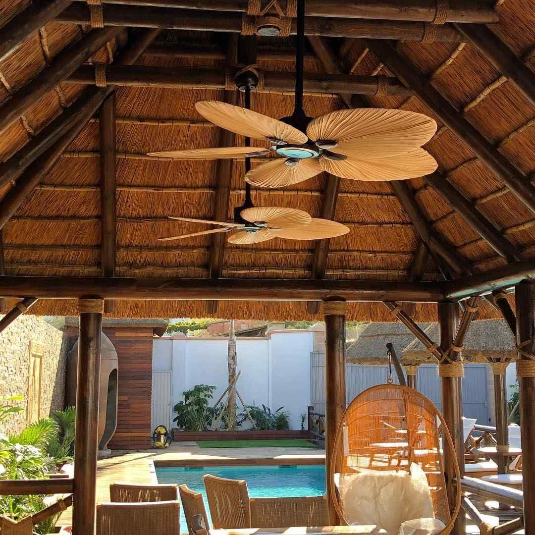 Cenador con techo de paja, ventiladores de techo y vista a la piscina. Cerca hay una silla colgante.