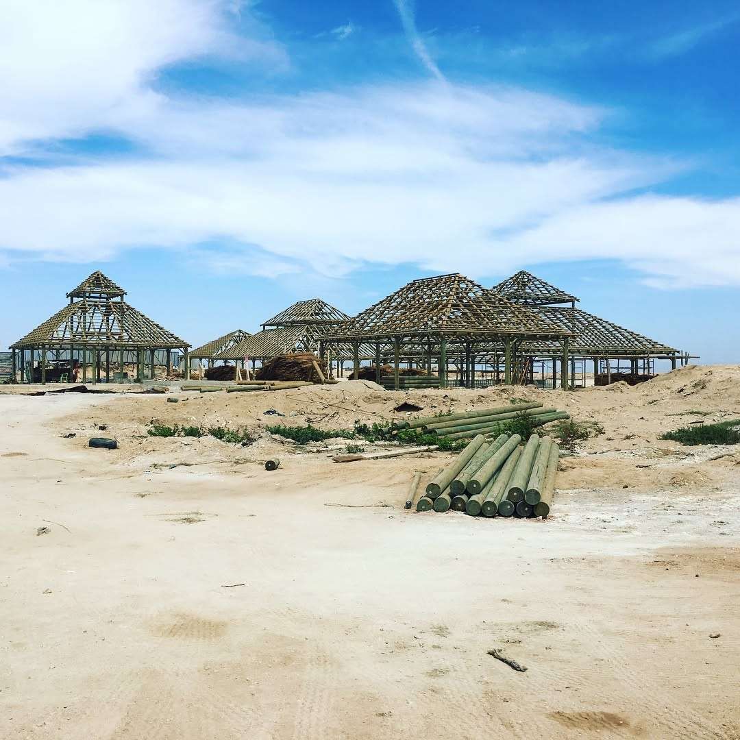 Construcción de varias estructuras de madera con techos de paja en una playa de arena bajo un cielo azul.