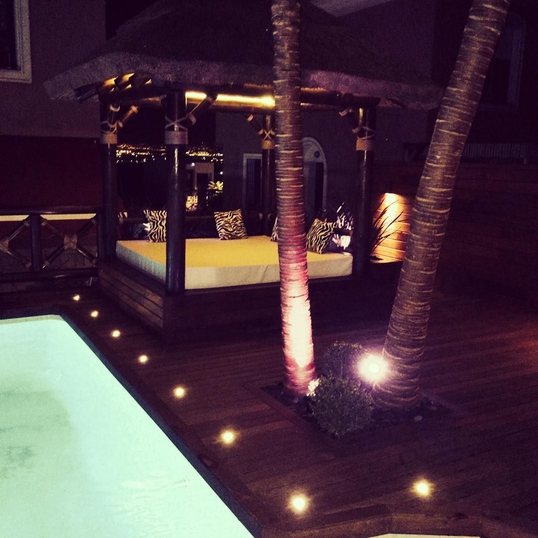 Escena nocturna: cenador con asientos junto a la piscina, iluminado por luces cálidas, palmeras.