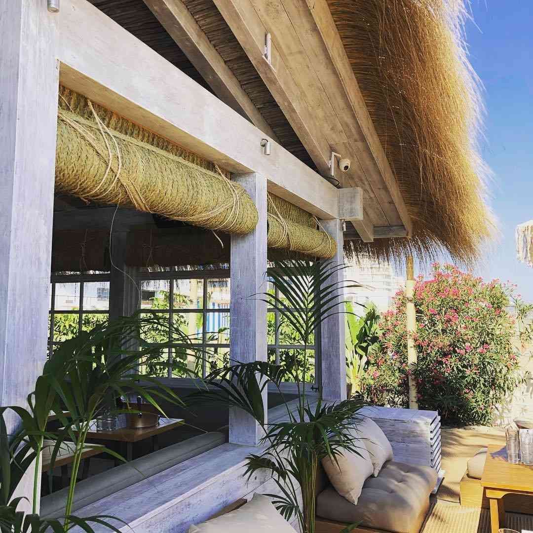 Cafetería frente a la playa con techo de paja, vigas blancas, cortinas tejidas y asientos al aire libre.