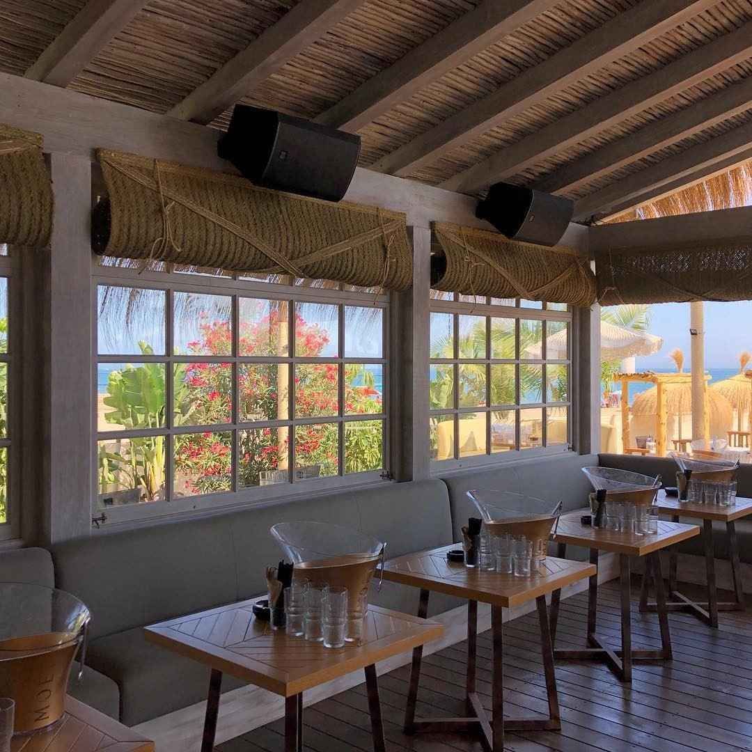 Restaurante interior junto a la playa con vistas. Mesas, bancos, ventanas y altavoces visibles.