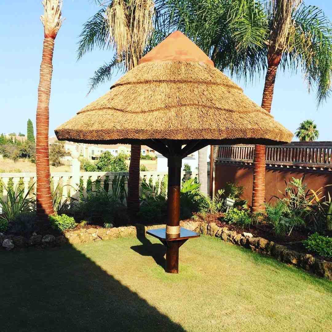 Cenador con techo de paja en un jardín soleado, con palmeras y un exuberante césped verde.