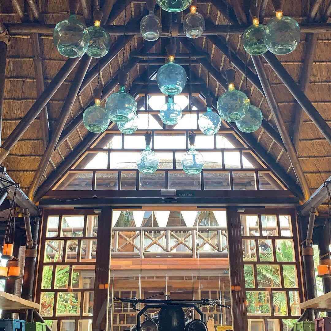 Vestíbulo con vigas de madera, techo de paja, grandes ventanales y lámparas de cristal colgantes.